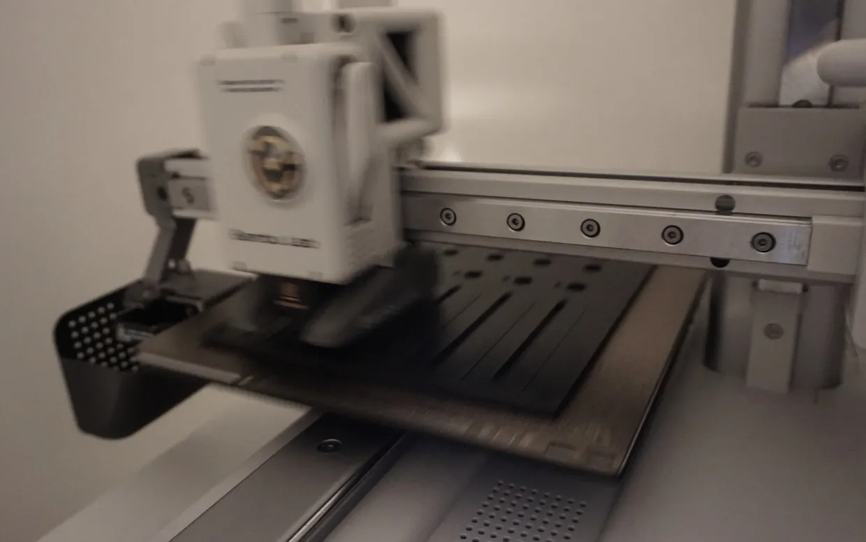 A white 3D printer at work, its print head blurred in motion over a black front panel. The slots for the faders and the holes for knobs and buttons are already visible in the partly printed plastic.