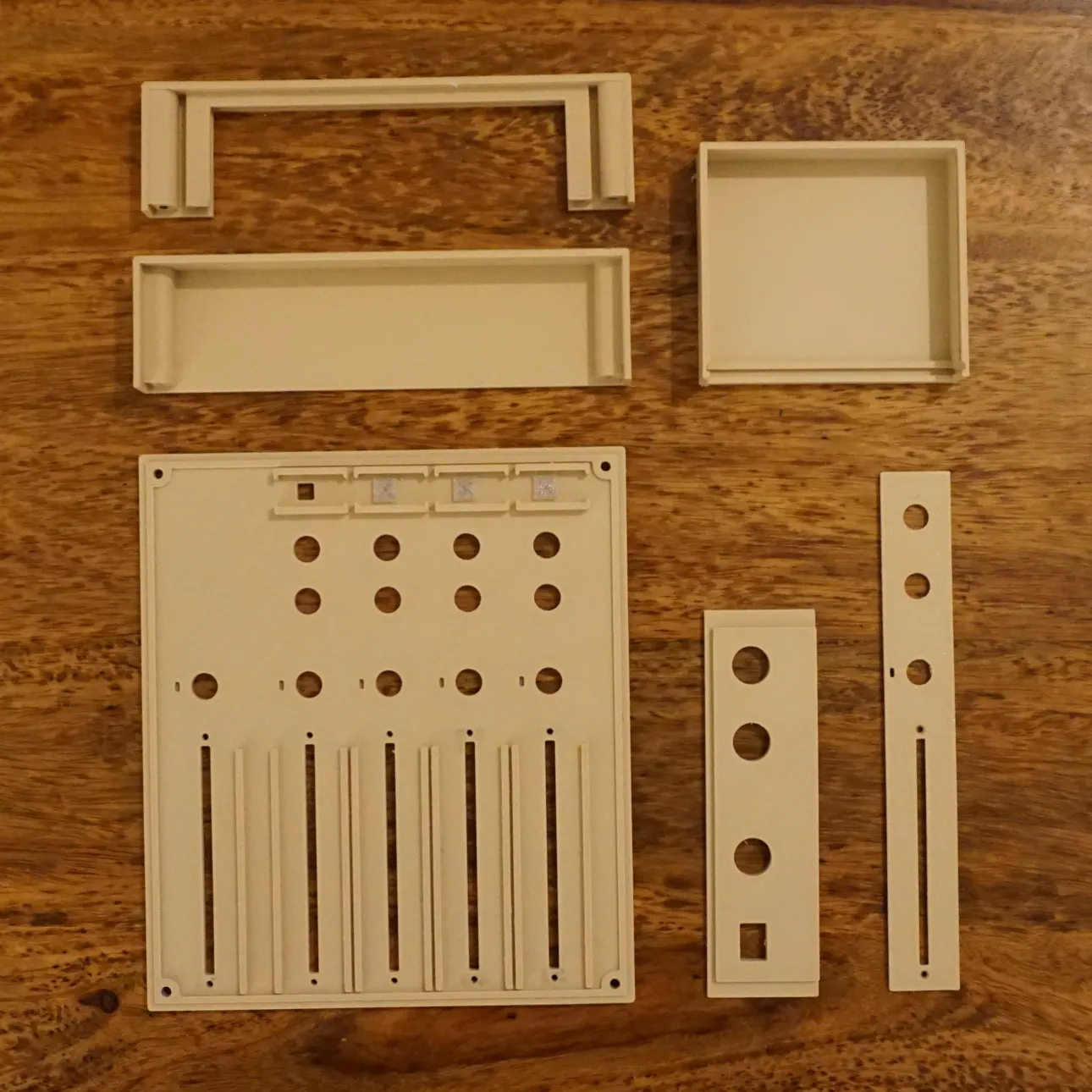 Beige test prints on a wooden desk: an early version of the full front panel with round holes and fader slots, two narrow test strips with holes and a fader slot, and a few box-shaped parts.