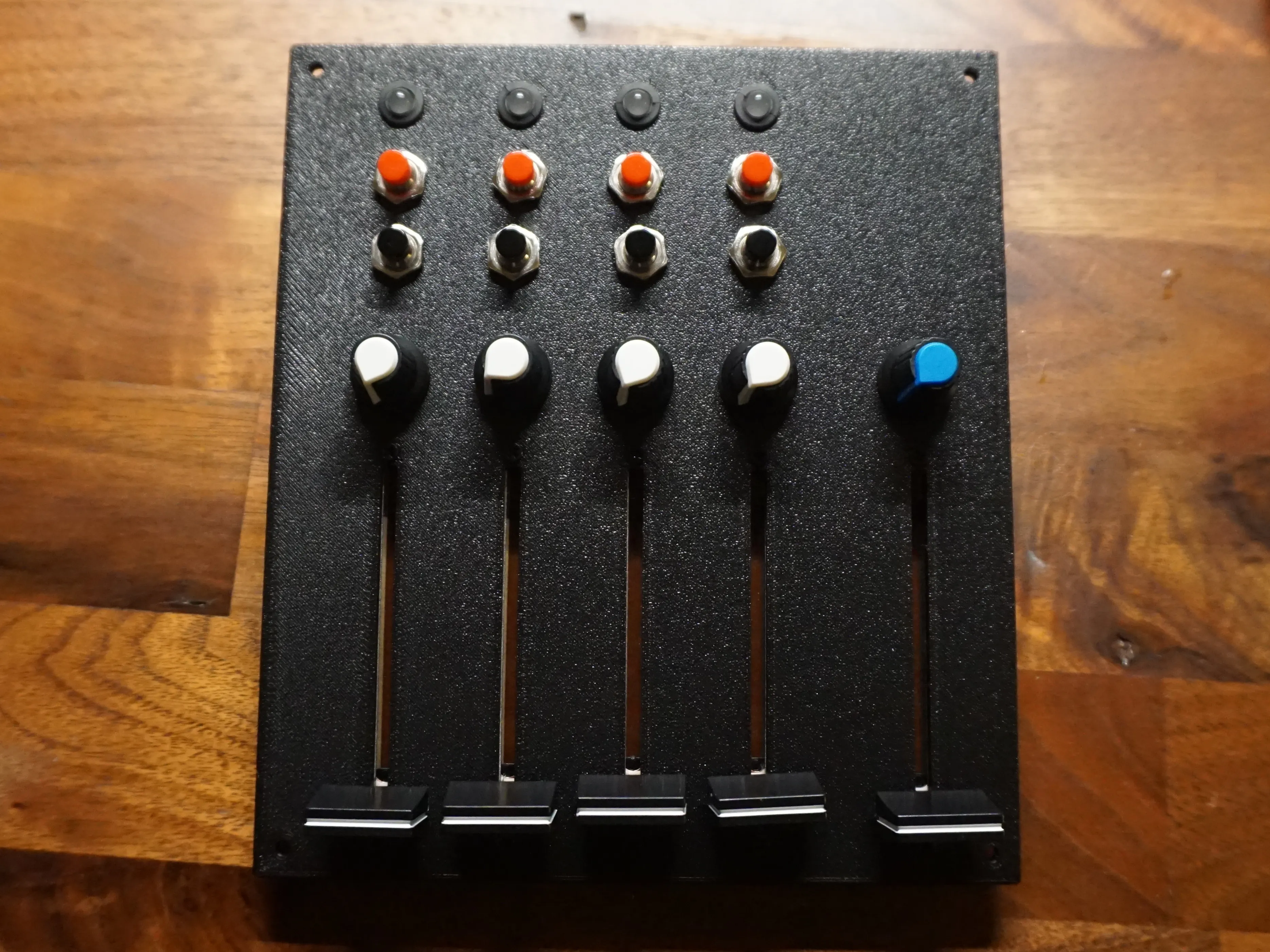 The printed black front panel on a wooden desk, with a slightly grainy, textured surface. Four columns each have an LED in a neoprene holder, a red and a black push button, a white knob and a long fader. A fifth column on the right has a blue knob and another fader.